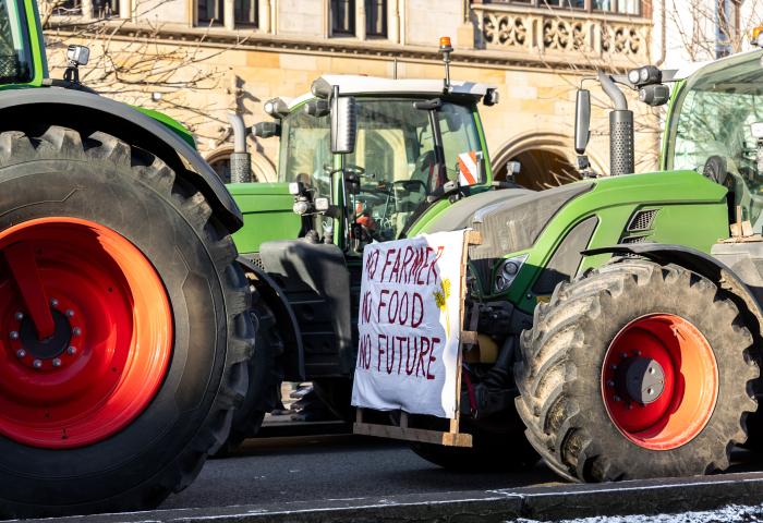 Grüne Traktoren auf Straße, an einem ein weißes Transparent mit rotem Text 'NO FARMER NO FOOD NO FUTURE'