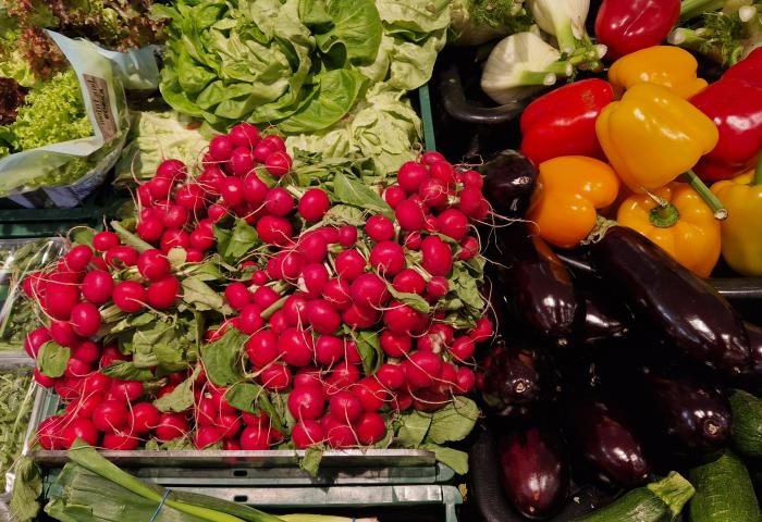 Frische, rote Radieschen mit grünen Blättern, daneben bunte Paprika in Gelb, Orange und Rot, Auberginen und Salatköpfe in einer Obst- und Gemüseabteilung.