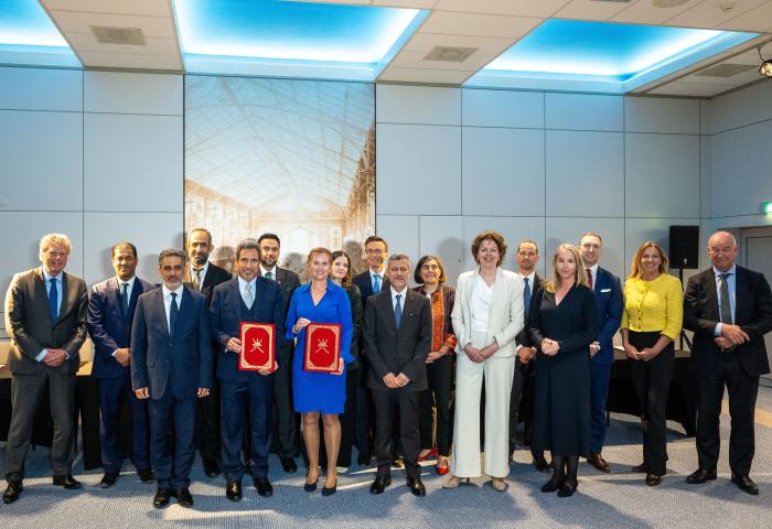 Gruppe von Personen in formeller Kleidung, zwei halten rote Ordner mit goldenen Emblemen, Hintergrund mit großem Bild einer Halle und blauer Deckenbeleuchtung.