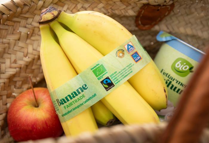 Bündel gelber Bananen mit Fairtrade-Etikett, daneben roter Apfel und Becher mit grünem Bio-Logo in geflochtenem Korb.