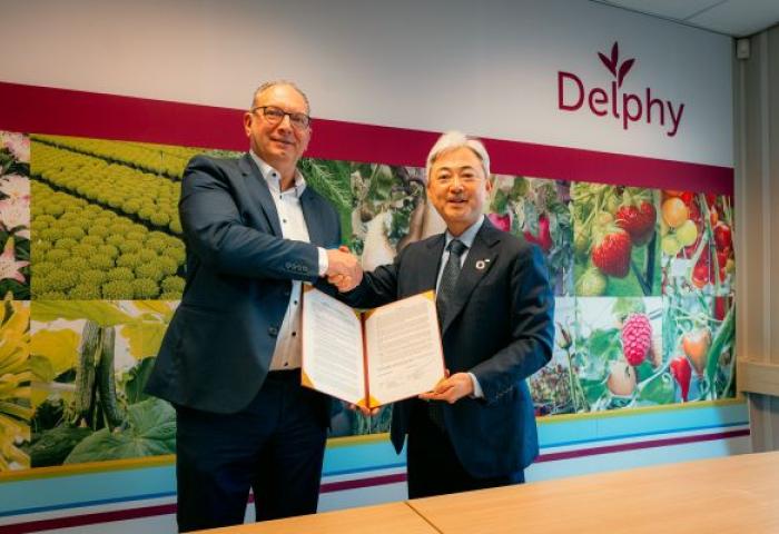 Zwei Männer in Anzügen schütteln sich die Hand, einer hält ein geöffnetes Dokument, im Hintergrund bunte Bilder von Obst und Gemüse und das Logo 'Delphy'.