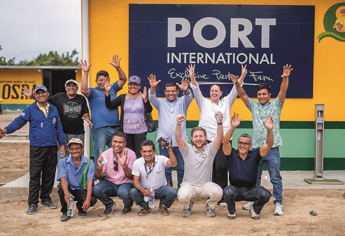 Gruppe von Menschen vor gelb-grünem Gebäude mit großem Schild 'PORT INTERNATIONAL Exclusive Partner Farm', einige mit erhobenen Händen.
