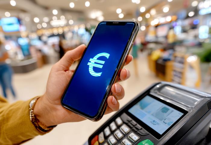 Ein in einer Hand gehaltenes Smartphone, welches ein Eurozeichen anzeigt, wird vor einem Bezahlterminal gehalten.