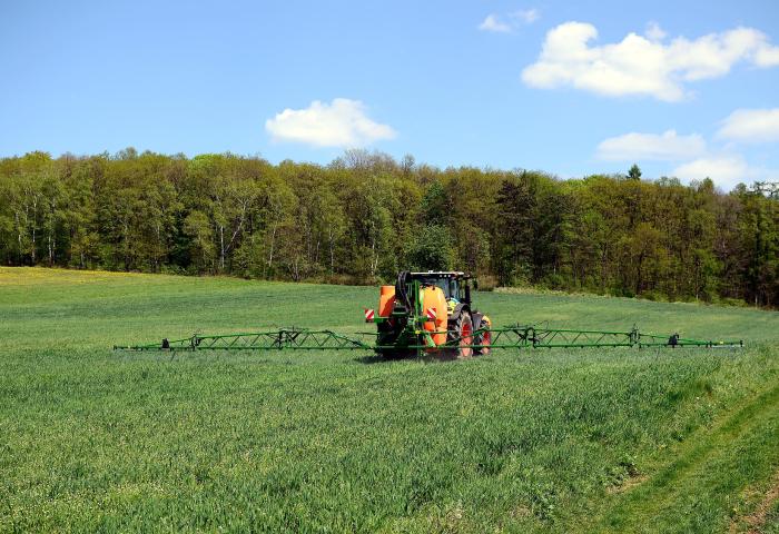Traktor mit breitem Pflanzenschutzgerät fährt über grünes Feld vor Wald und blauem Himmel mit Wolken.
