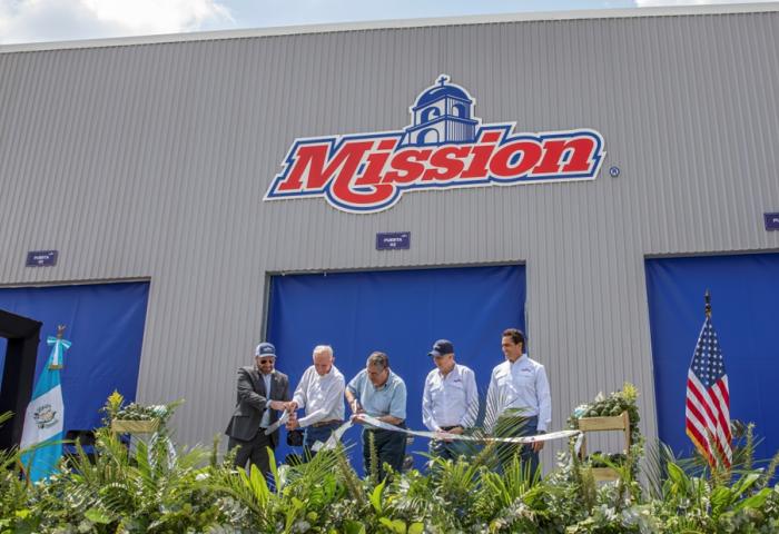 Fünf Männer stehen vor einem Gebäude mit dem Schriftzug 'Mission' und halten ein Band, umgeben von Pflanzen, guatemaltekischer und US-amerikanischer Flagge.