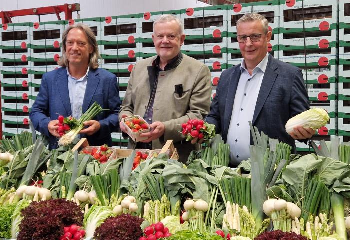 Christian Deyerling (l.), Hans-Jörg Friedrich (Bildmitte) und Reinhard Oerther stehen hinter einem Tisch mit vielfältigem frischem Gemüse, darunter Spargel, Radieschen, Salat und Chinakohl. Im Hintergrund sind gestapelte Kisten mit Pfalzmarkt-Logo zu sehen.