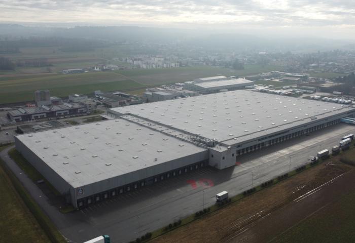 Luftaufnahme eines großen Industriegebäudes mit flachem Dach und mehreren runden Dachaufbauten, umgeben von Feldern und einer Stadt im Hintergrund.
