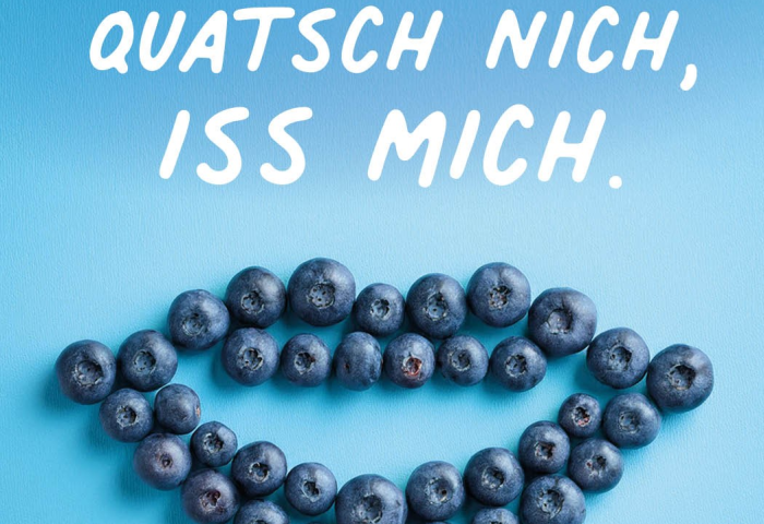 Mehrere Heidelbeeren bilden die Form eines Mundes auf blauem Hintergrund, darüber der Text 'QUATSCH NICH, ISS MICH.' in weißer Schrift.