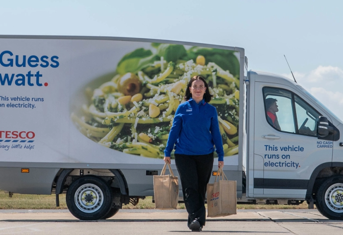 Eine Lieferwagen von Tesco auf dem Rollfeld eines Flughafens. Davor trägt eine Tescomitarbeiterin lächelnd  zwei Einkaufstaschen.
