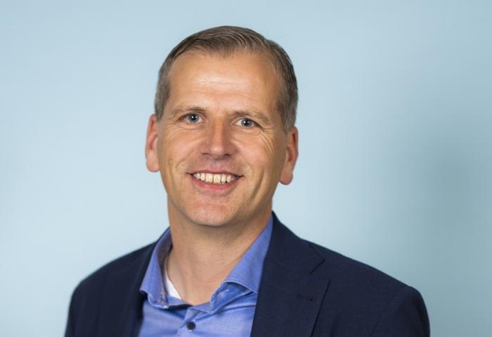Mann mit blonden Haaren, dunkelblauer Jacke und hellblauem Hemd.