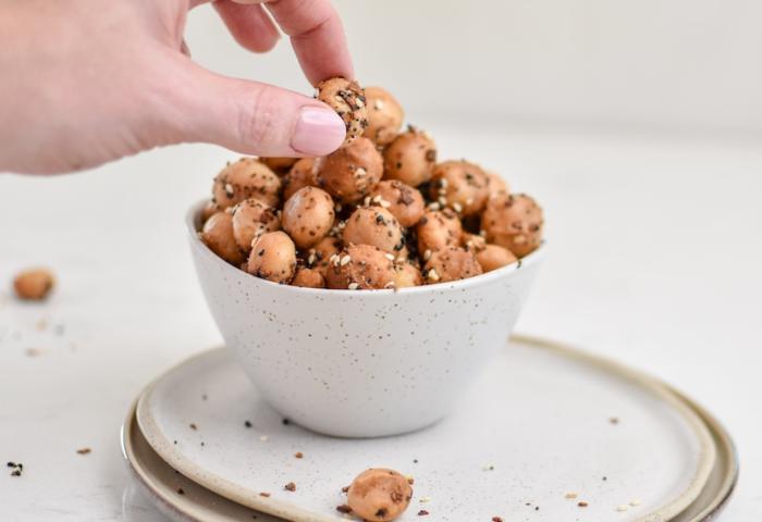 Mit Gewürzen versehene Macadamias in einer weißen Schlüssel. Eine weibliche Hand greift zu.