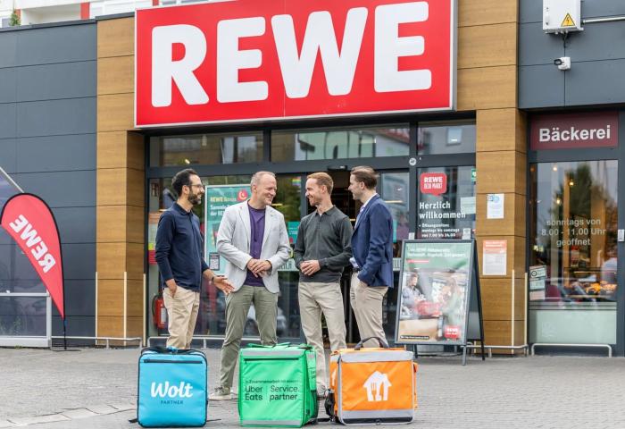 vier Männer stehen mit Transportboxen vor einer Rewe-Filiale