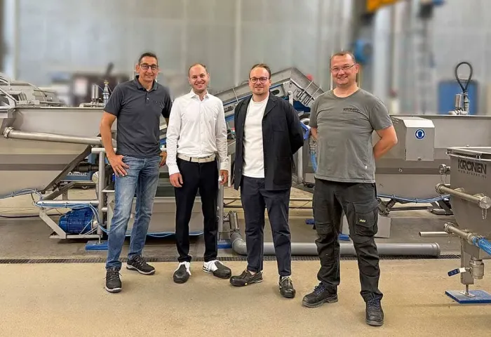 Vier Männer stehen vor einer KRONEN-Salatverarbeitungslinie mit Edelstahlmaschinen und Förderbändern in einer Produktionshalle.
