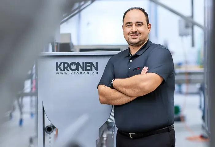 Wissam Badr, Leiter Finanzen und Prokurist der KRONEN GmbH, steht mit verschränkten Armen neben einer Maschine mit KRONEN-Logo.