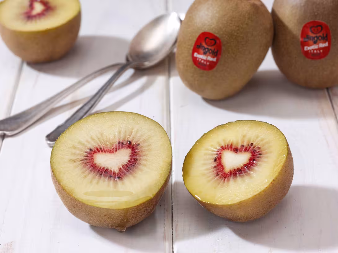 Zwei Hälften einer aufgeschnittenen Jingold Exotic Red Kiwi mit rotem Inneren, auf einem weißen Tisch. Im Hintergrund sind ganze Kiwis und zwei Löffel sichtbar.